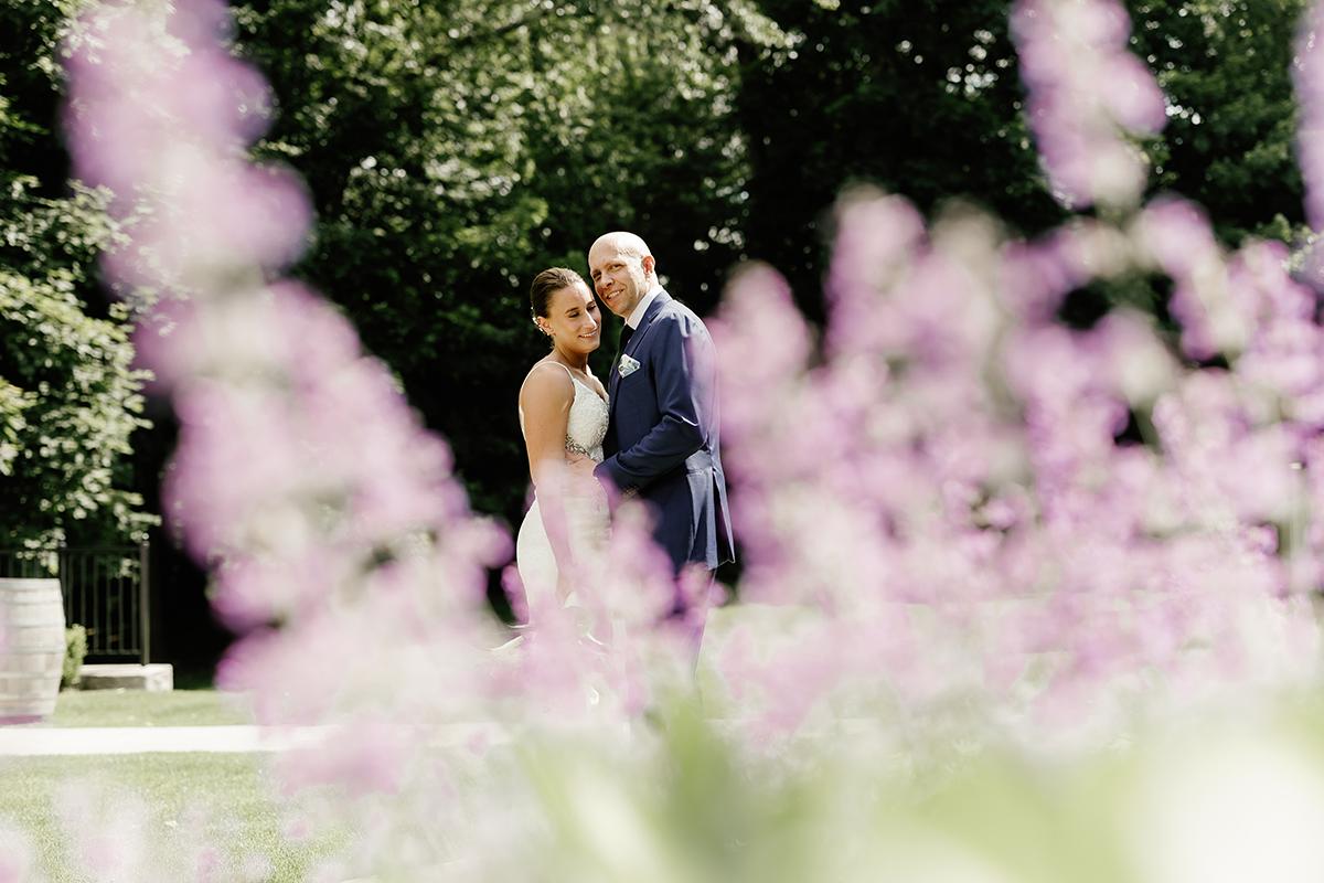 Bride and groom embrace, blurred lavender flowers in foreground, lush trees behind.