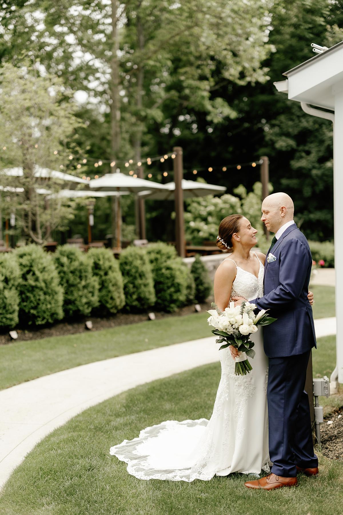 Bride and groom stand smiling in a garden, surrounded by greenery and lights.