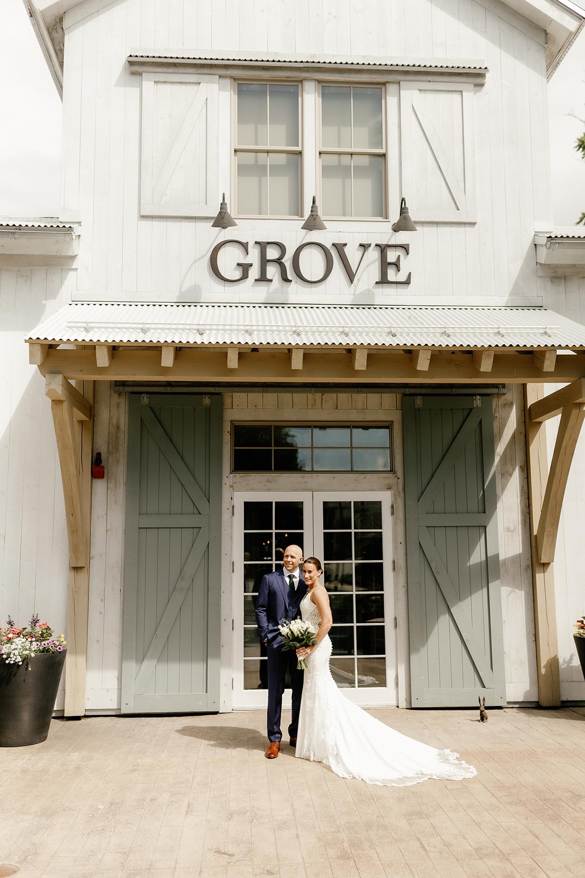 Bride and groom standing in front of a rustic white building with "GROVE" sign.