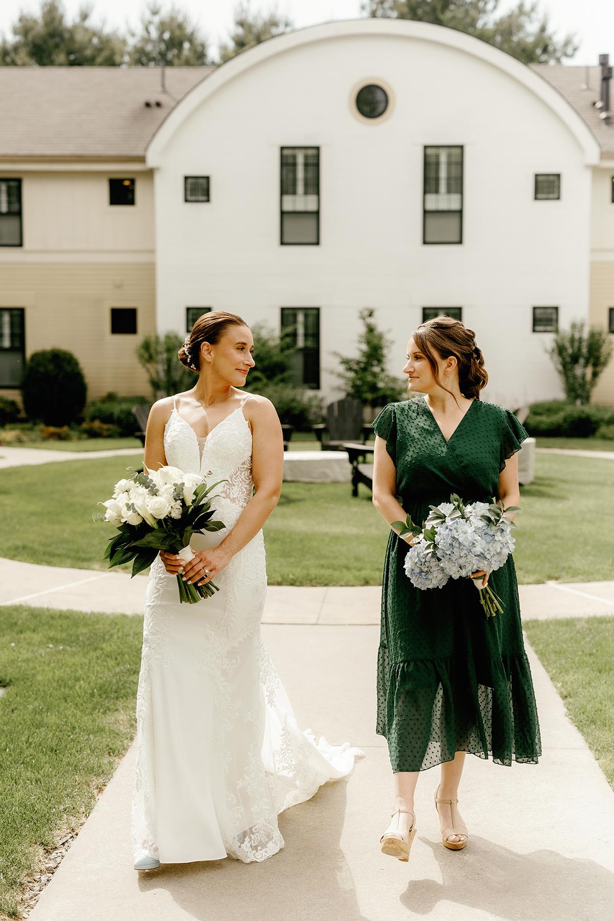 Bride and bridesmaid walking on a path, holding bouquets, smiling at each other.