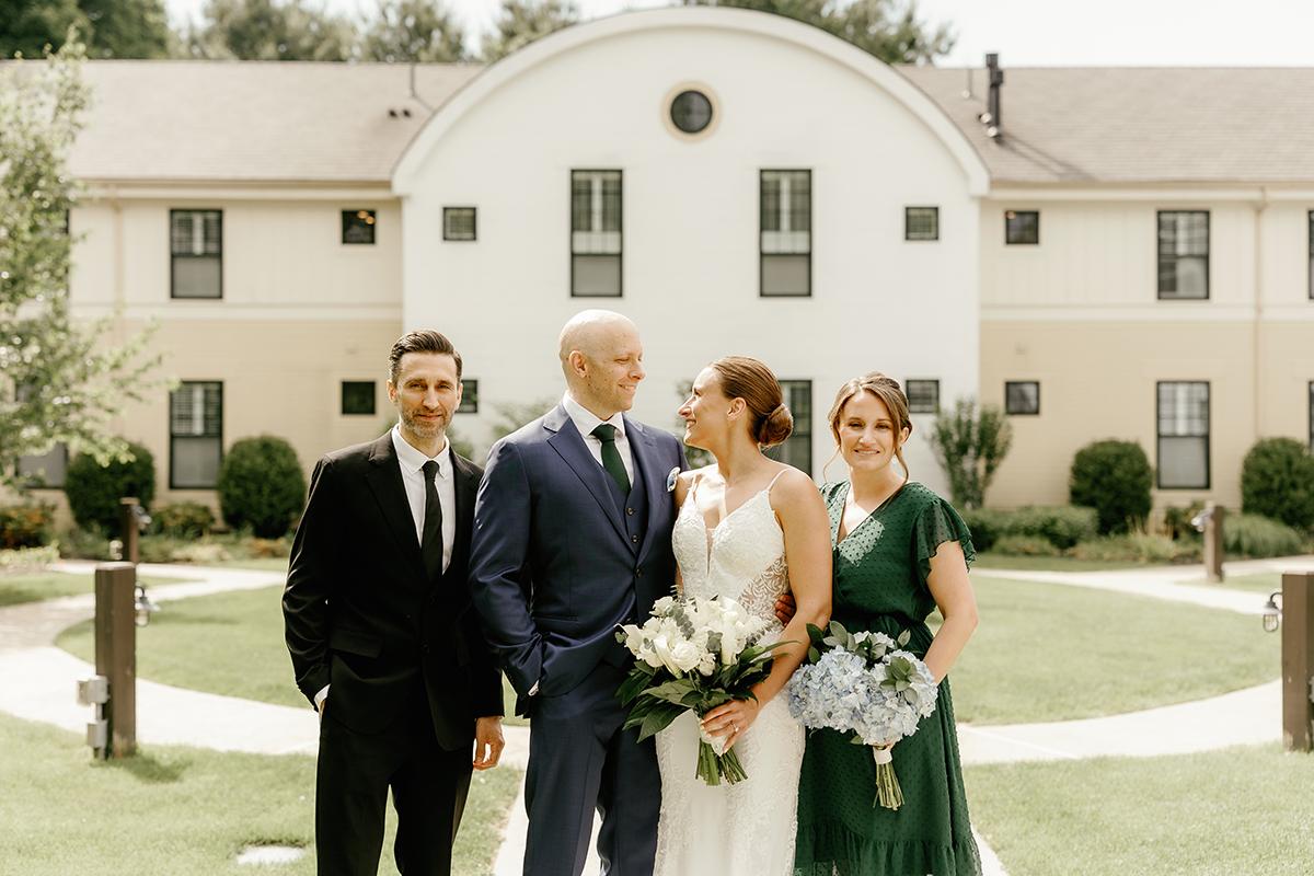 Wedding party standing in front of a white building, smiling, with bouquets.