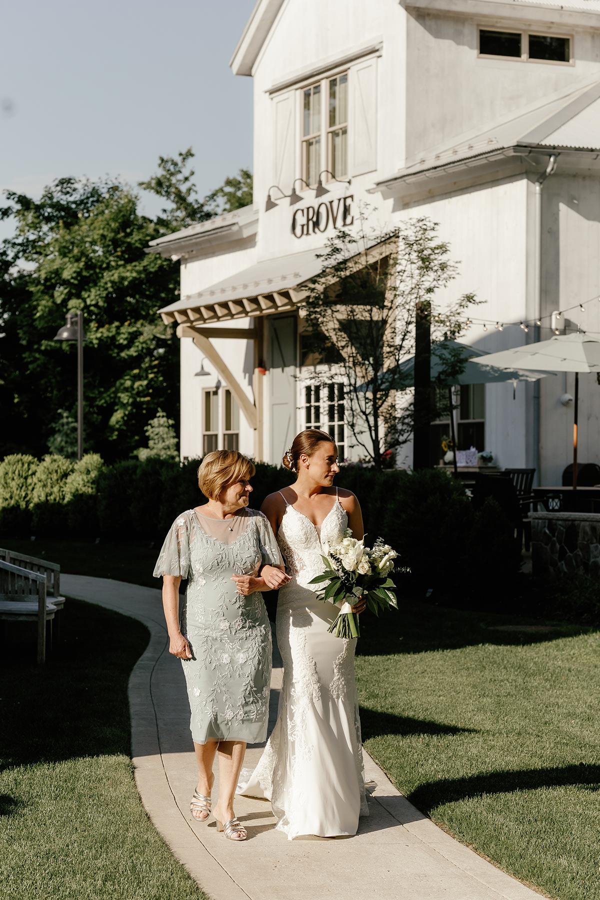 Bride with bouquet walks with woman in blue dress on sunny path near a white building.