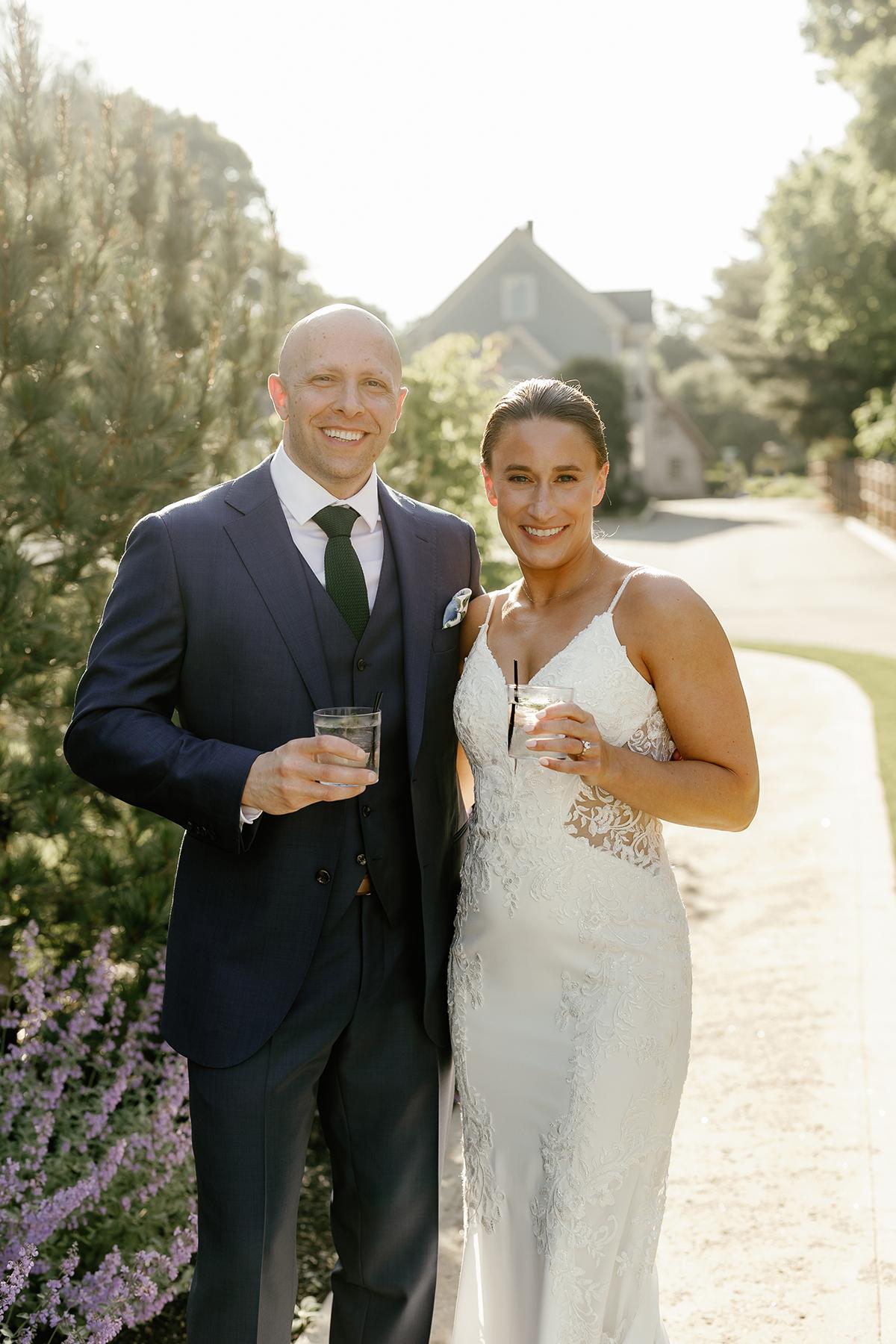 Couple in wedding attire smiling outdoors, holding drinks.