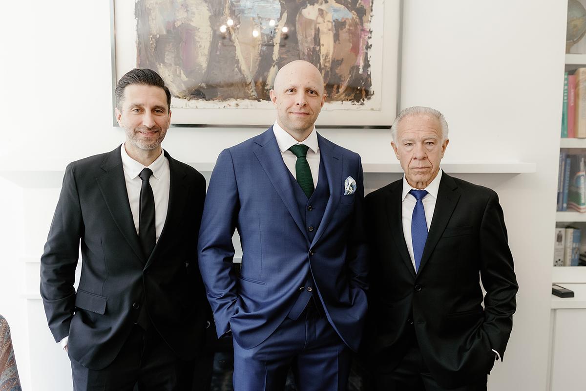 Three men in suits stand together; one in blue, two in black, in a room with art and books.