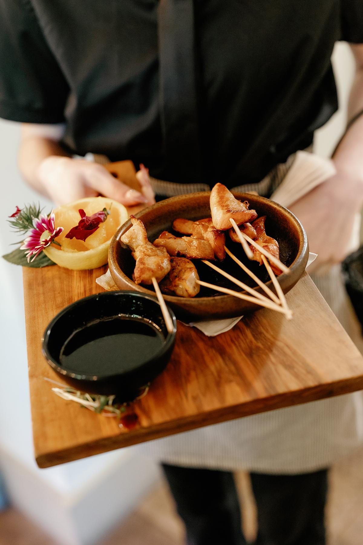 Server holding skewered appetizers with sauce and garnish on a wooden tray.