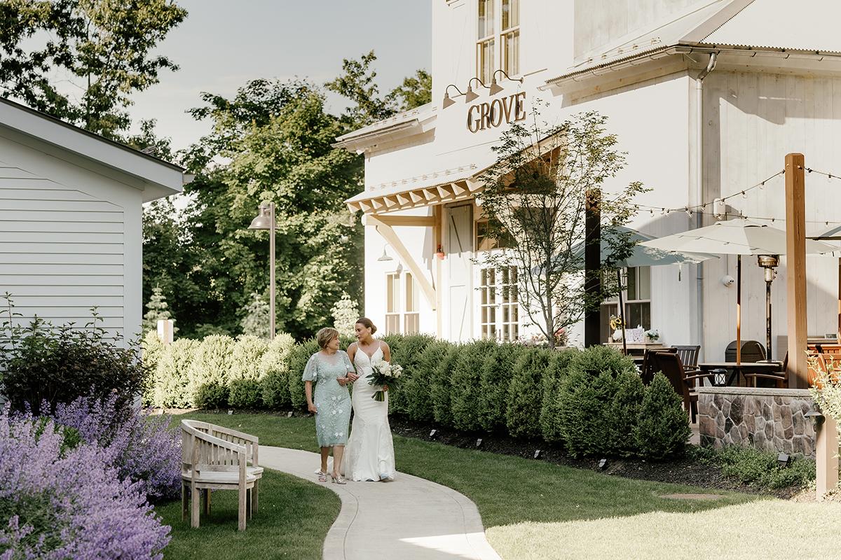 Bride and woman walking along garden path near a white house.