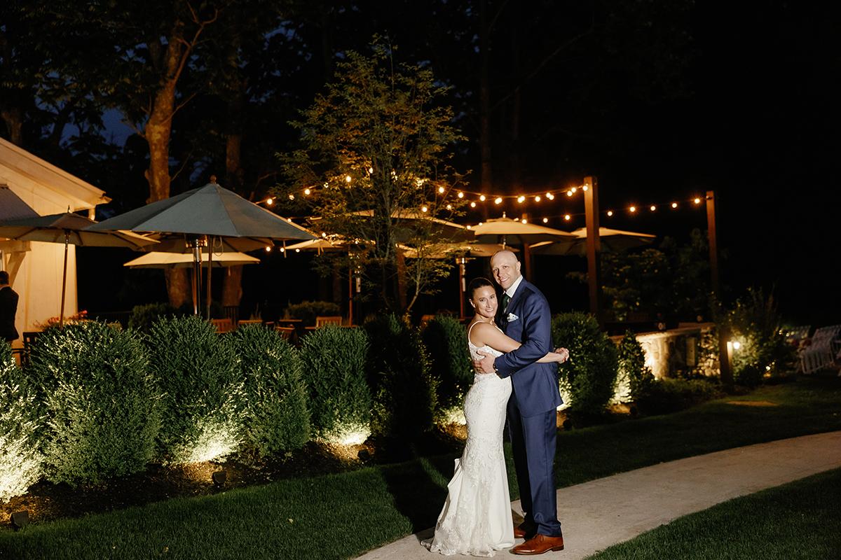 Bride and groom embrace on a lit garden path at night.