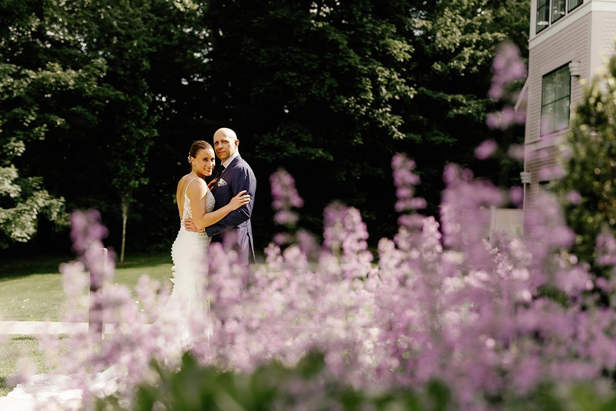 Bride and groom embrace in garden with purple flowers in foreground.