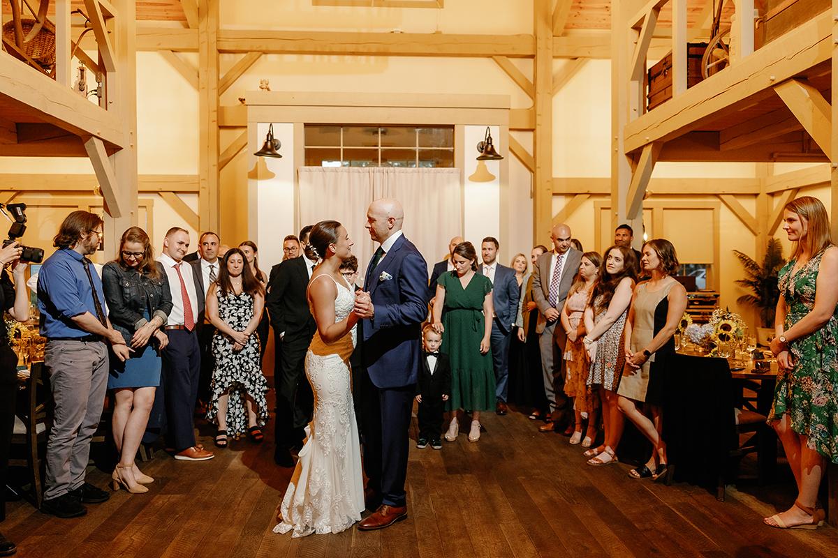 Bride and groom dance surrounded by guests in a warmly lit hall.