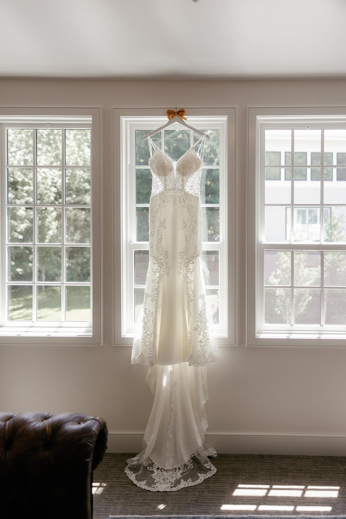 White wedding dress hanging in bright window, sunlight streaming in.