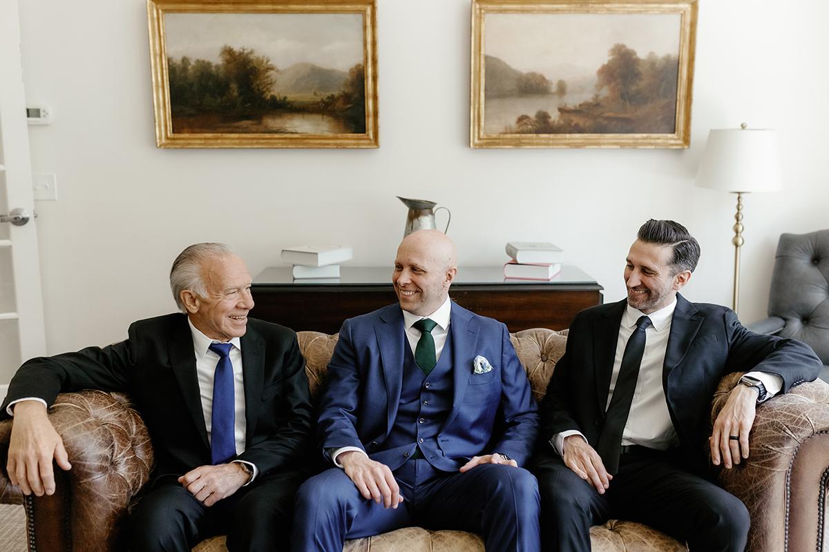 Three men in suits sitting on a brown sofa, smiling and talking.