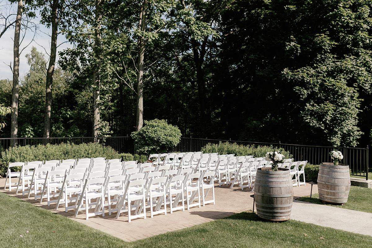 Outdoor wedding setup with white chairs and two barrels, surrounded by trees.
