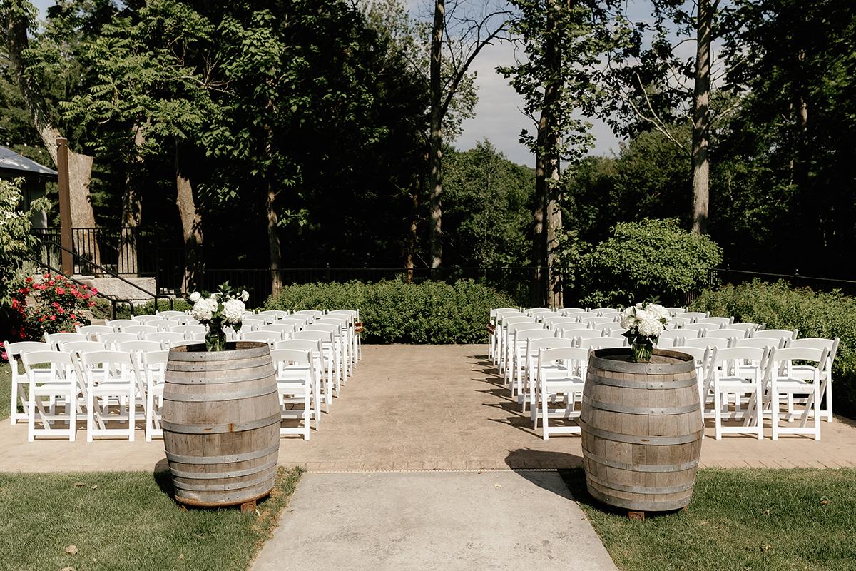 Outdoor wedding setup with white chairs, barrels, and trees in the background.