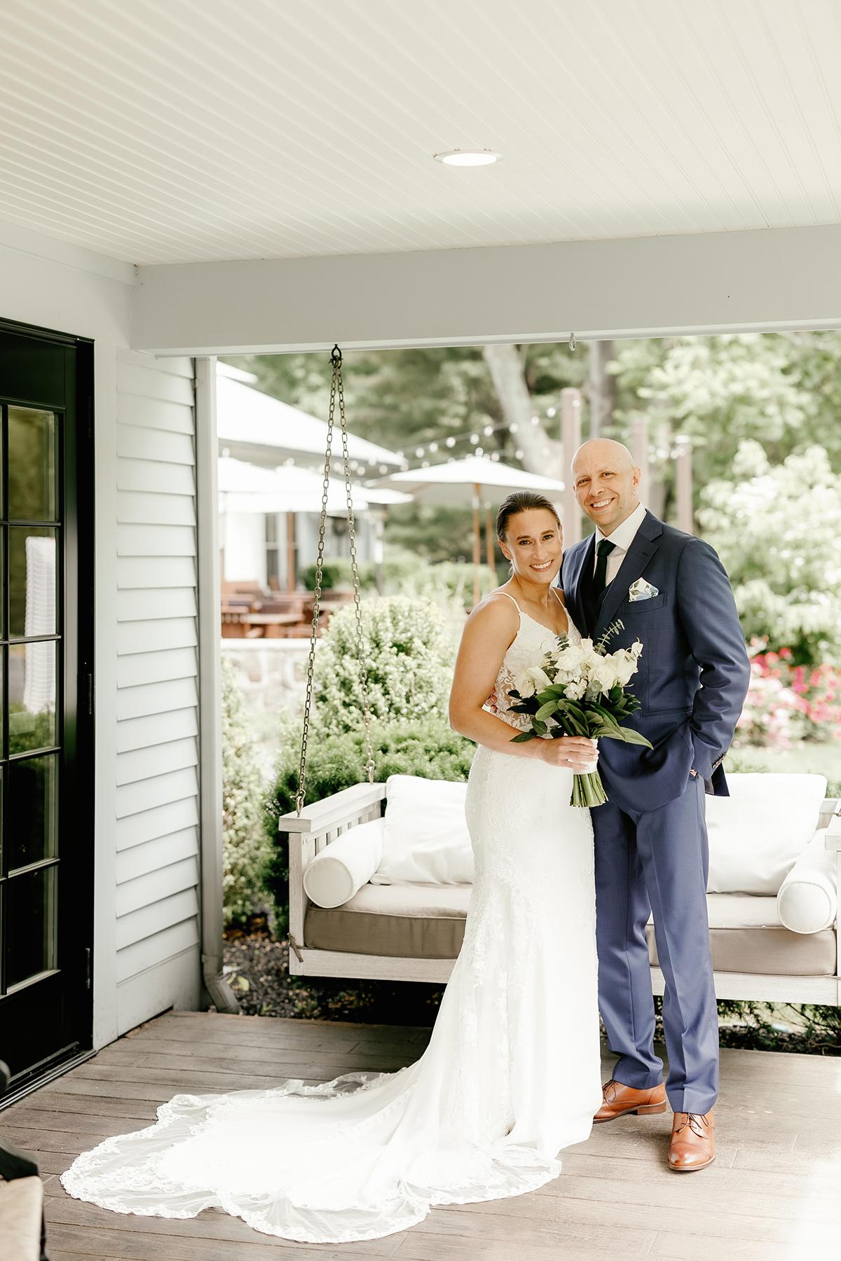 Bride and groom standing on a porch, smiling, with greenery in the background.