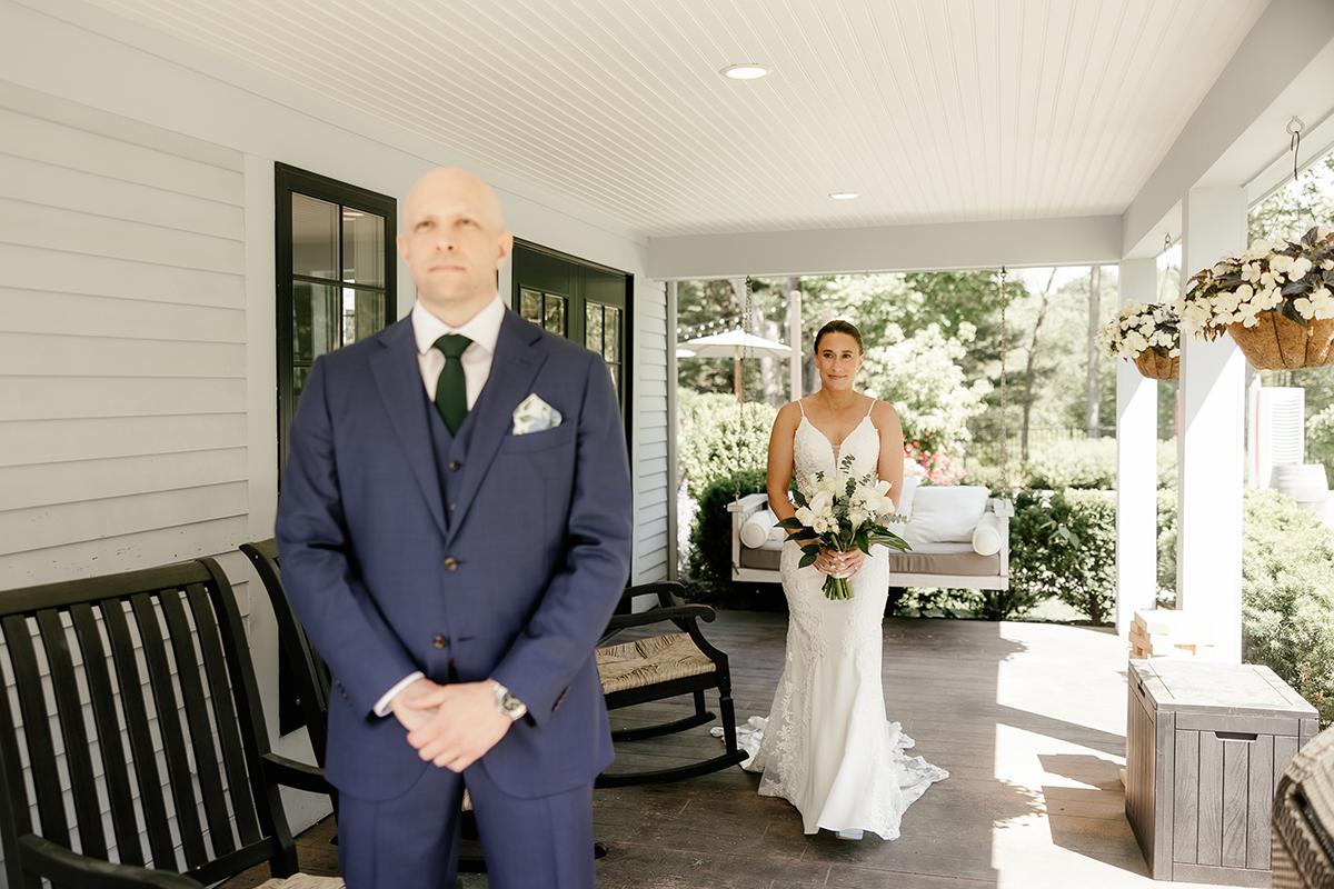 Bride approaches groom from behind on a sunlit porch.
