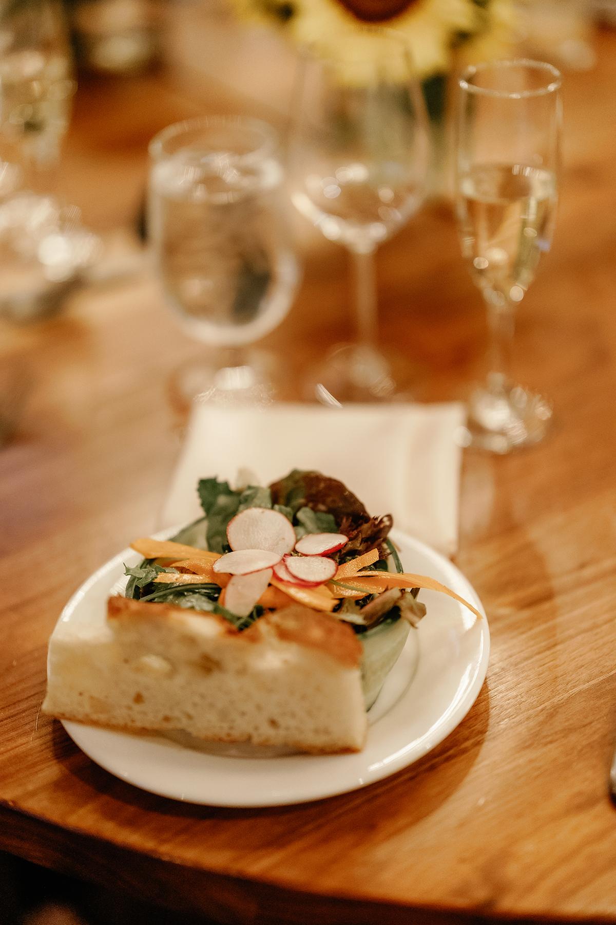 Focaccia topped with greens and radishes, on a wooden table with glasses in the background.