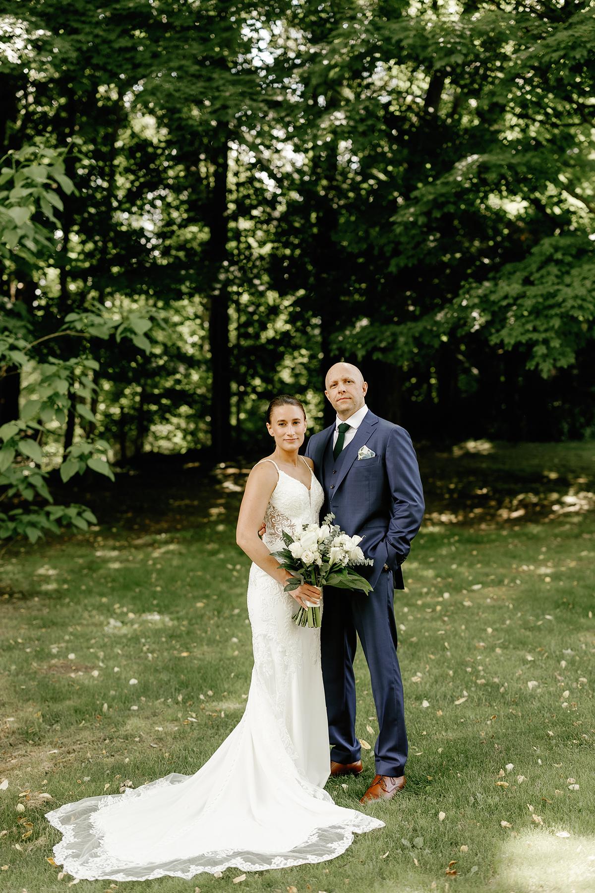 Bride and groom standing in a sunny forest, bride holding bouquet.
