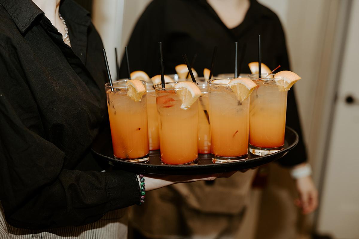 Tray with orange cocktails, garnished with lemon slices and straws, carried by server in black.