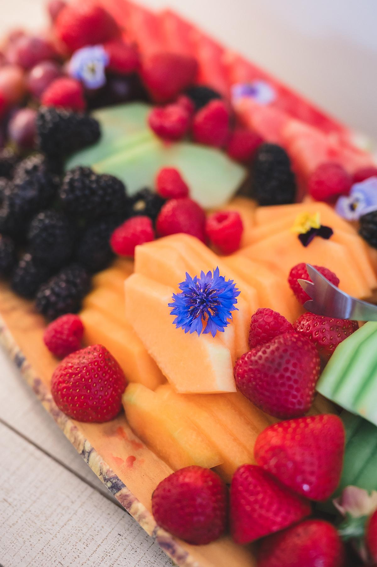 Sliced fruit platter with strawberries, melon, blackberries, and a blue flower.