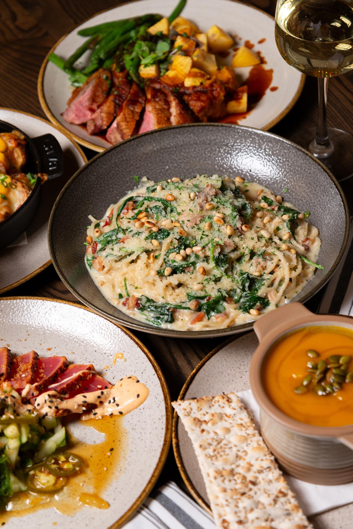 A variety of gourmet dishes including pasta, soup, and meat on a table with white wine.