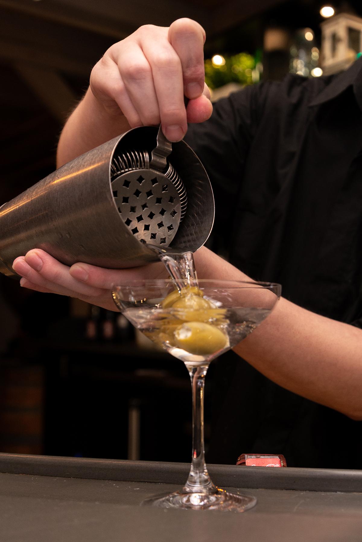 Bartender pouring martini into glass with olives.