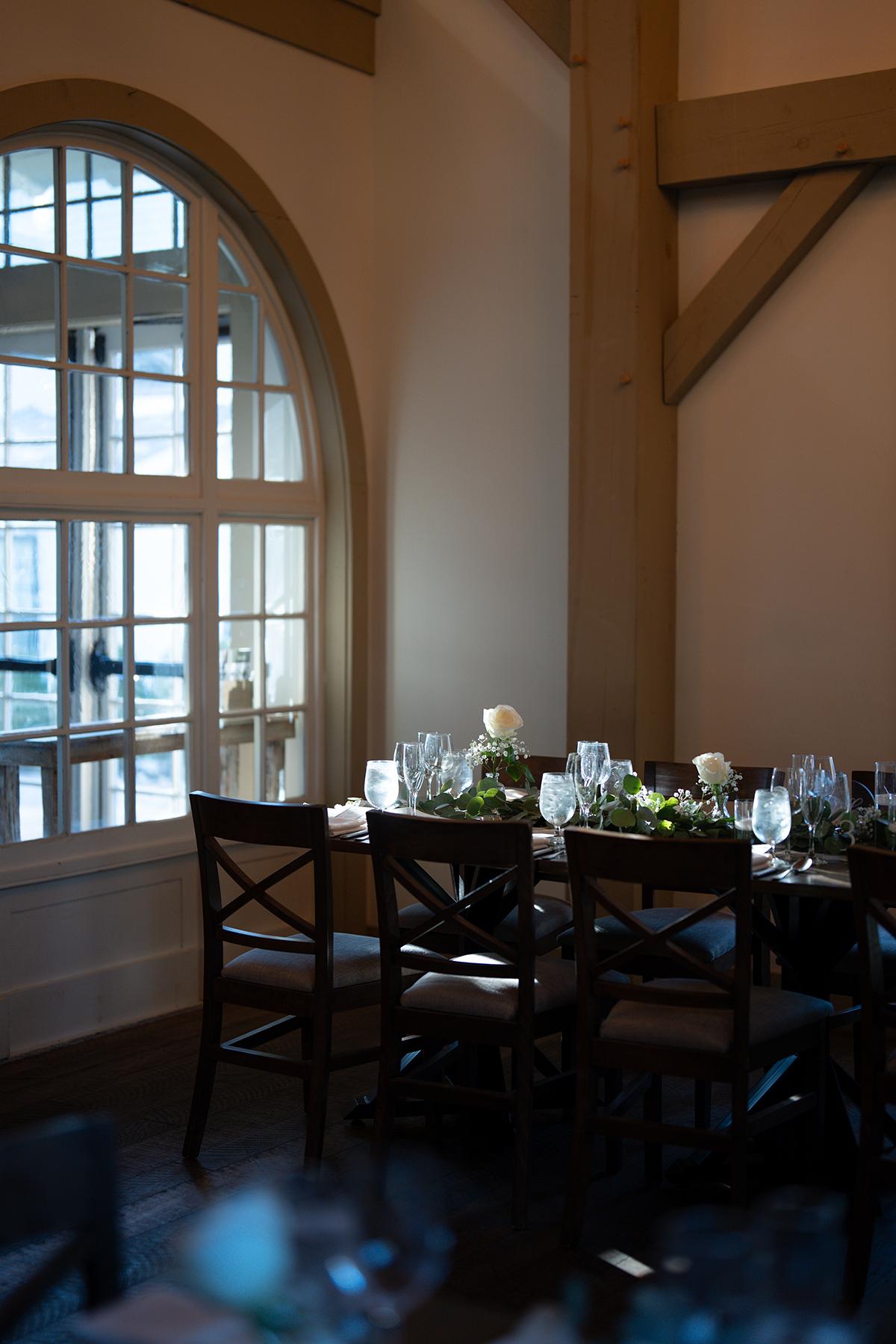 Dimly lit dining table with white roses and glassware by an arched window.
