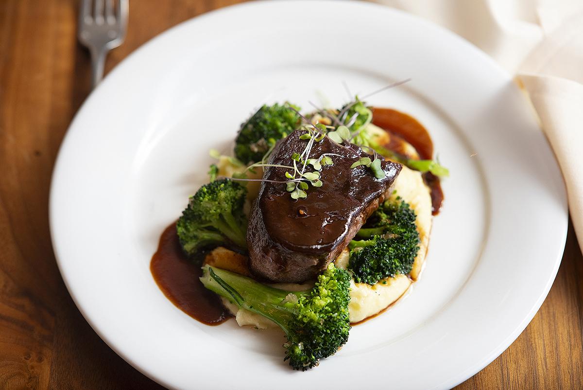 Steak with sauce, broccoli, and sprouts on a white plate.
