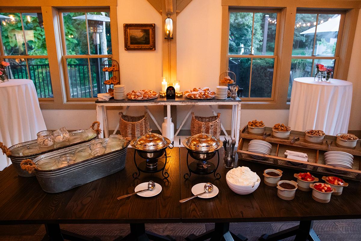 Buffet table with dishes, condiments, and drinks in a cozy room with large windows.