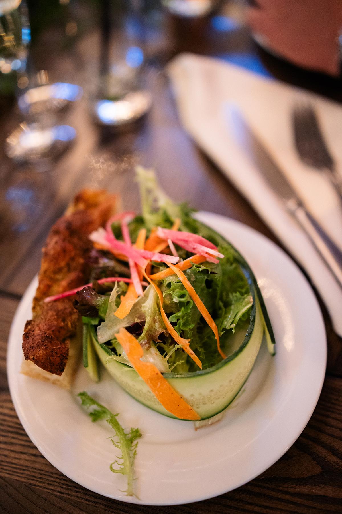 Salad wrapped in cucumber with a quiche slice on a white plate, set on a wooden table.