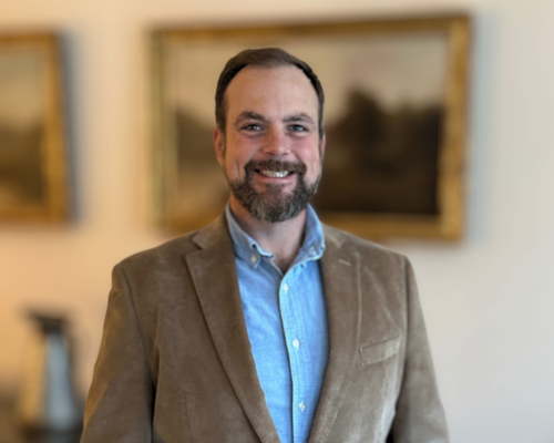 Smiling man in a brown blazer and blue shirt indoors.