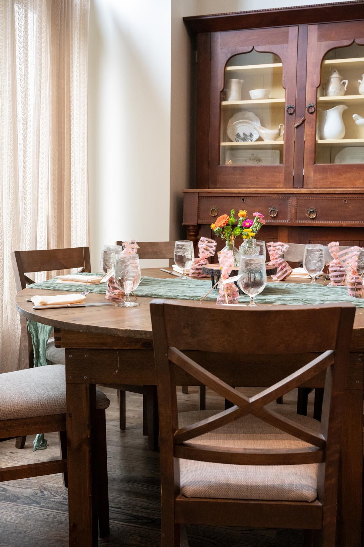 Dining table set with glassware, flowers, and a wooden cabinet in the background.