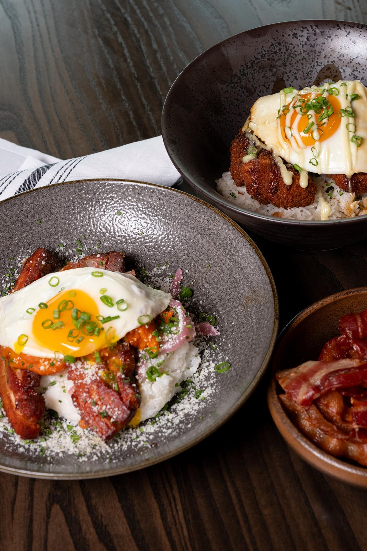 Two plates with fried eggs over sausages and rice, and a bowl of bacon, on a wooden table.