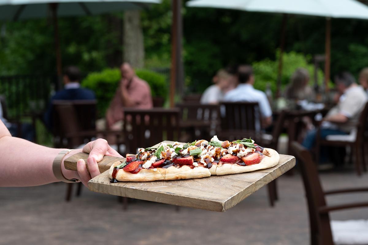 Pizza on wooden board outdoors with diners in background.