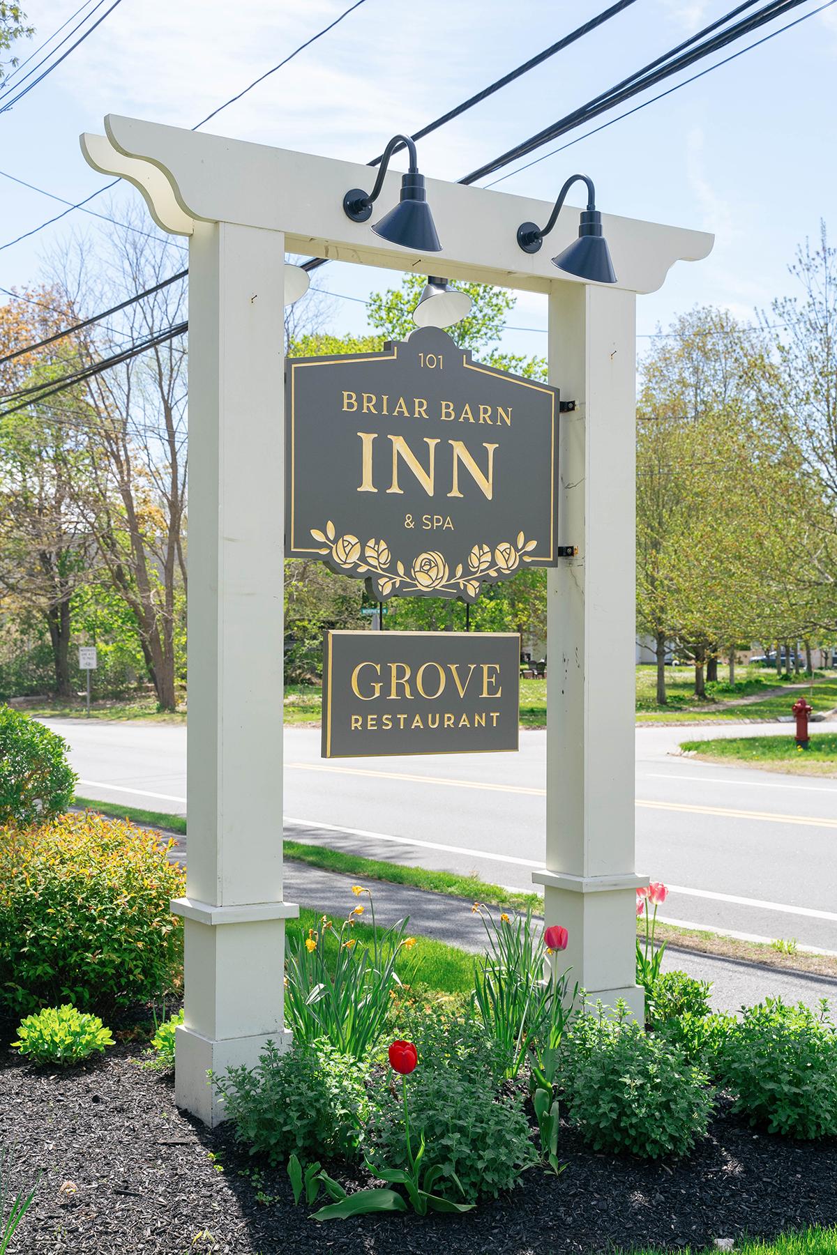 Inn sign with floral design, surrounded by greenery, on roadside.