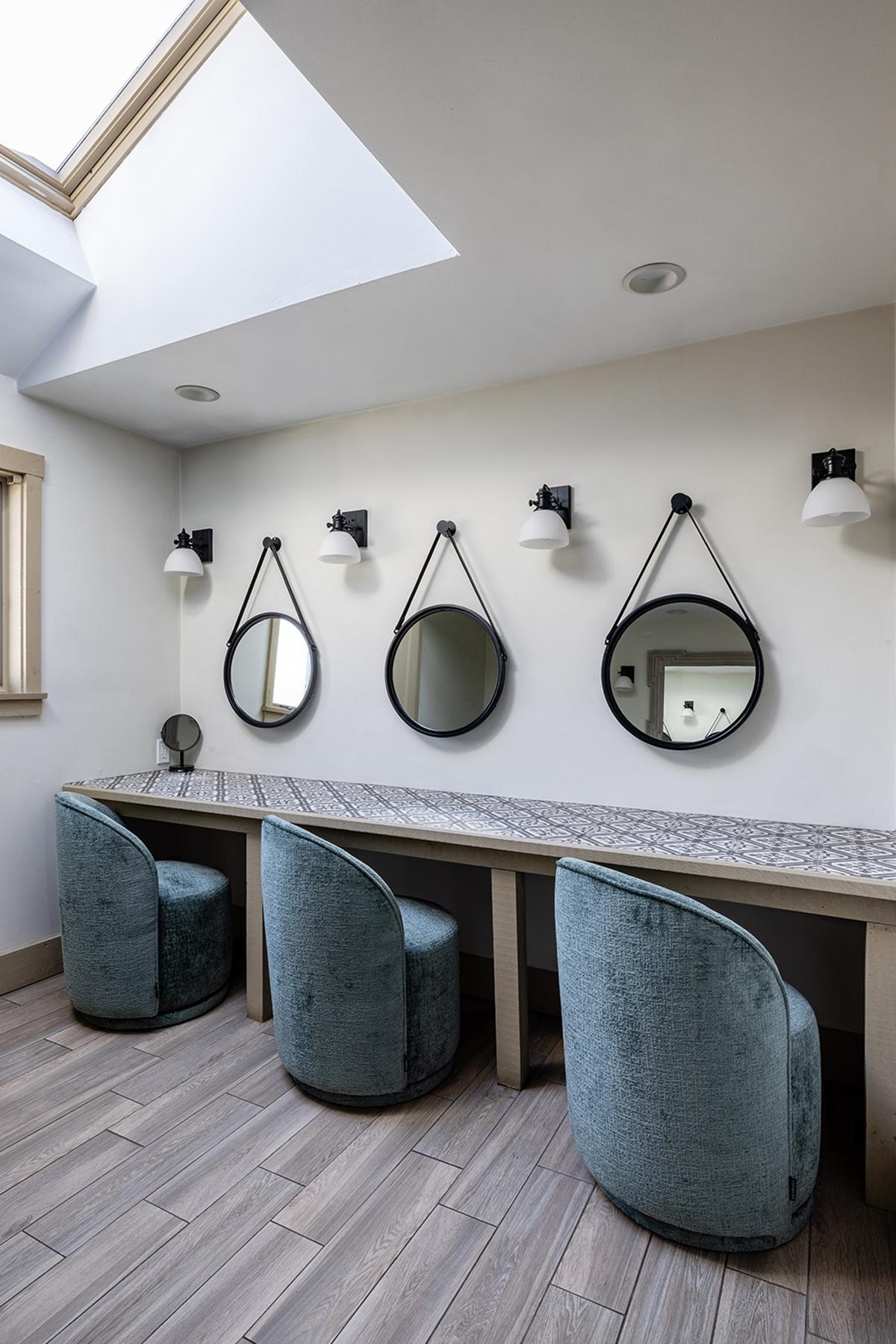 Modern vanity area with round mirrors, soft blue chairs, and skylight.