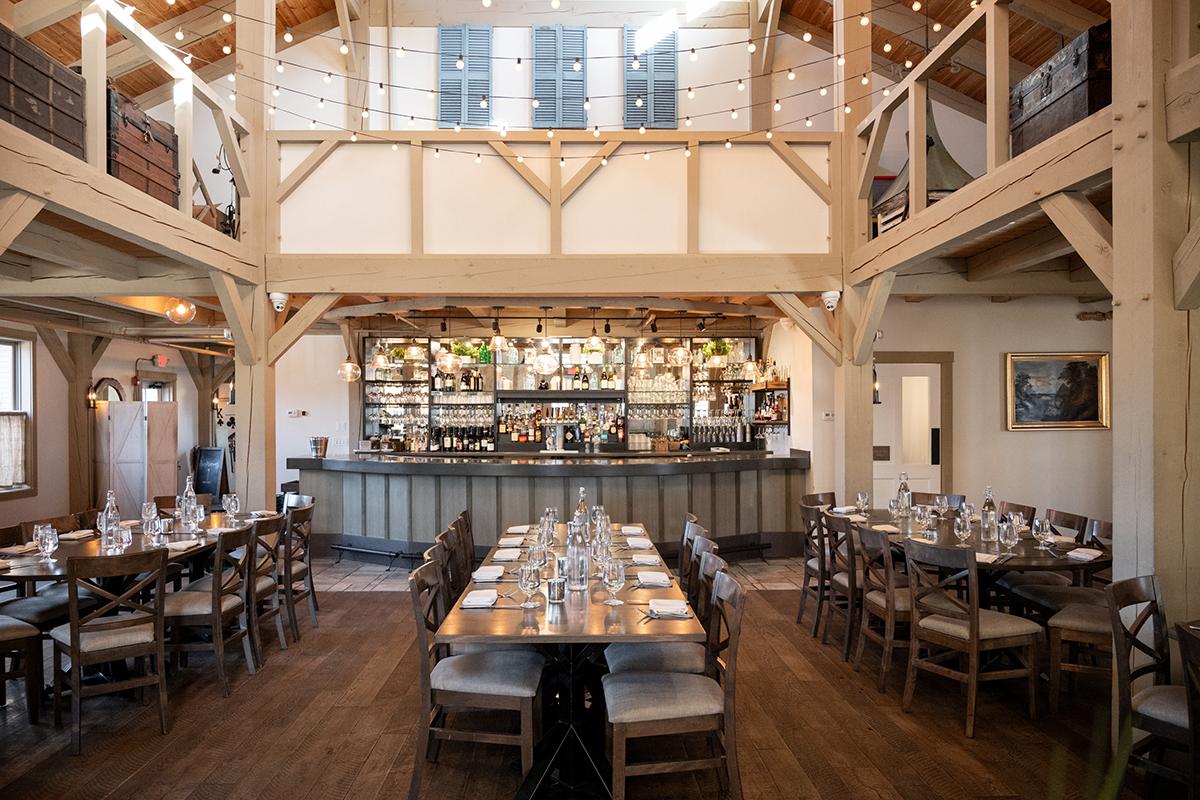Rustic restaurant interior with wooden tables and chairs, exposed beams, and a stocked bar.