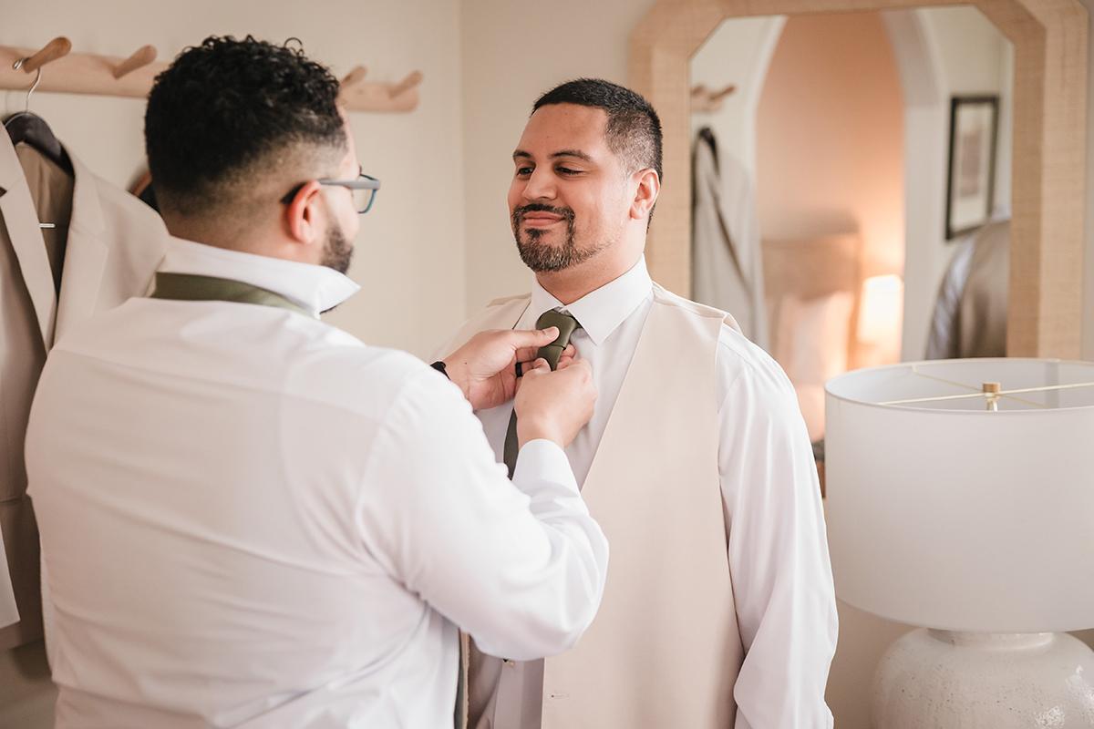 Man helps another adjust his tie in a warmly lit room.