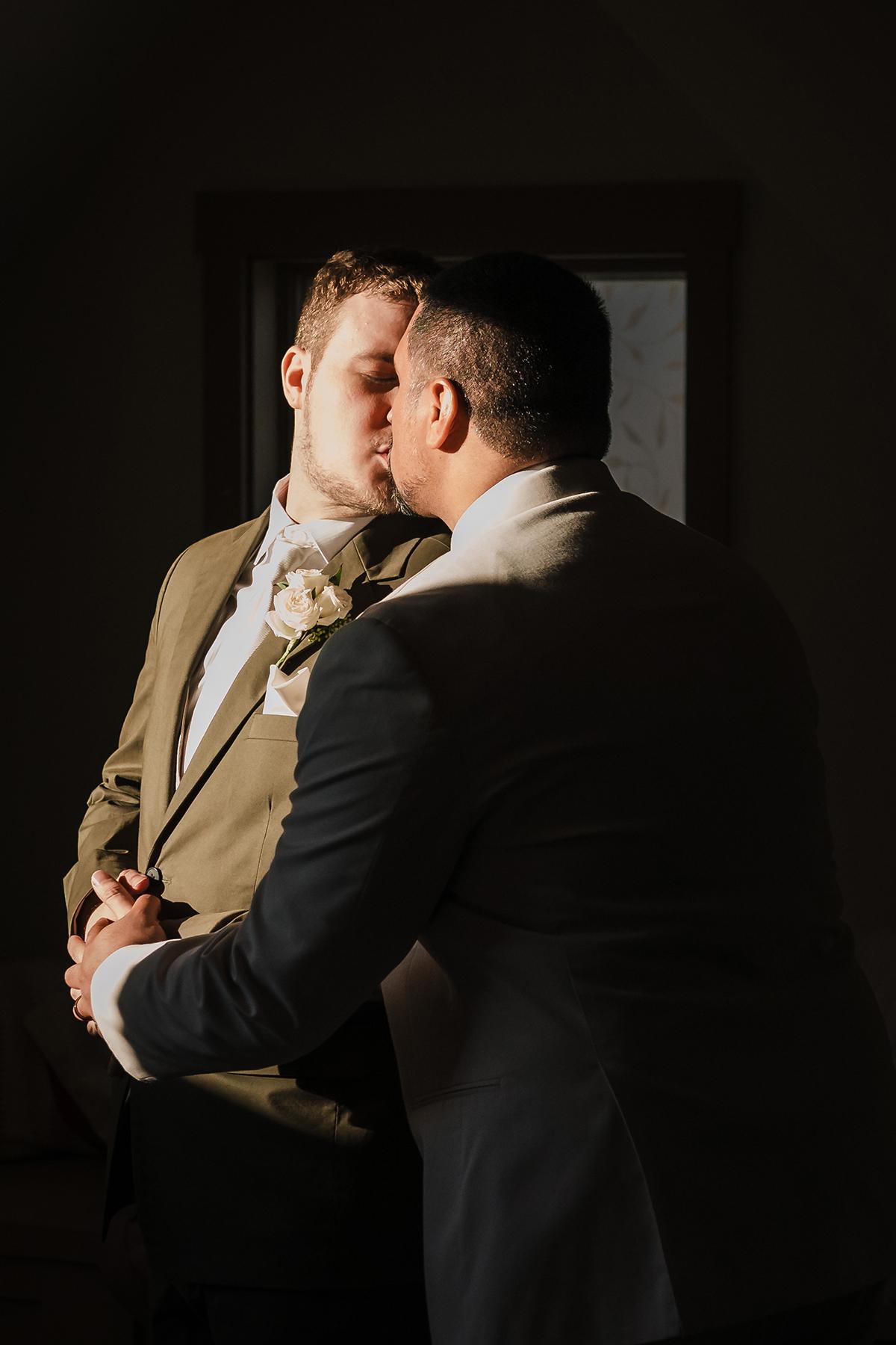 Two men in suits kissing in warm light.