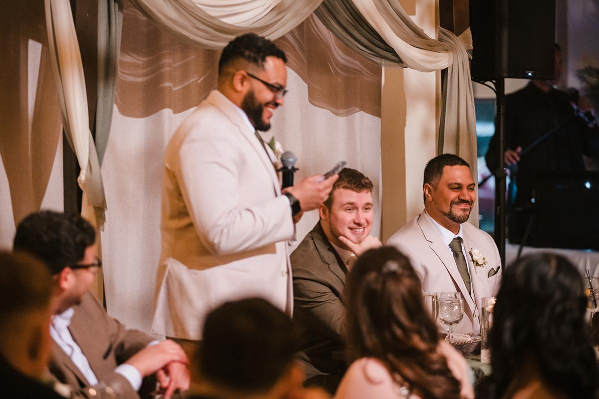 Man in white suit giving a speech, guests seated around, festive setting.