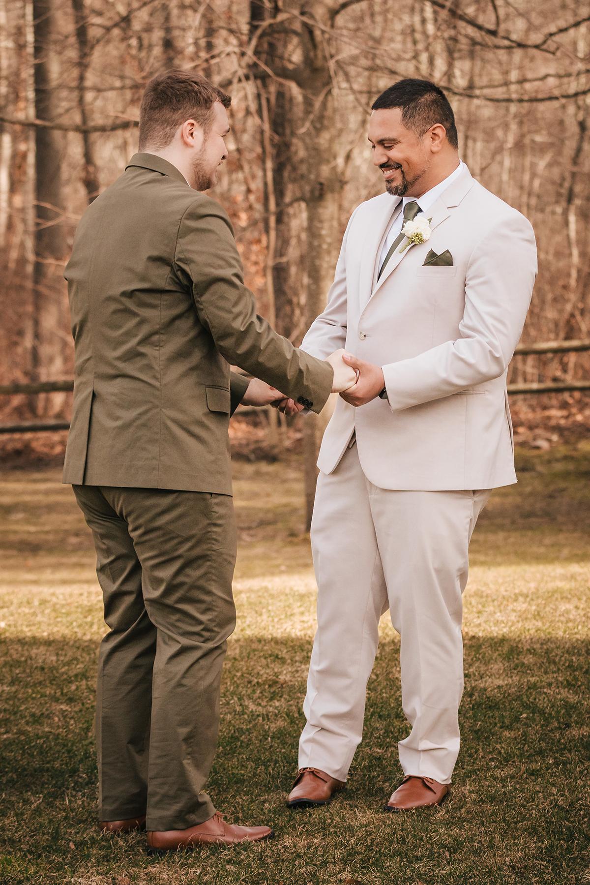 Two men in suits smiling, standing hand in hand outdoors.