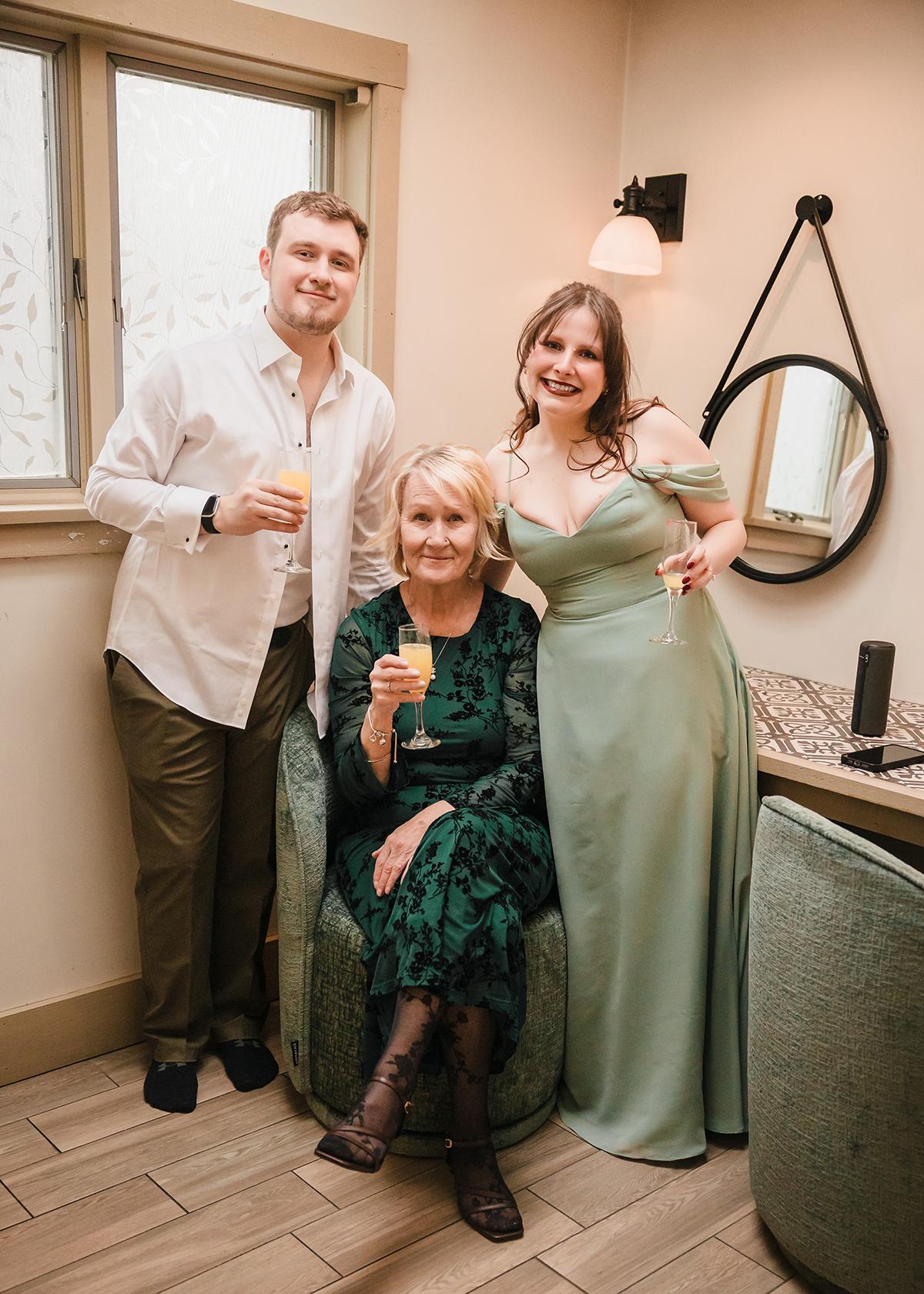 Man and woman stand beside seated older woman, all smiling, holding drinks.