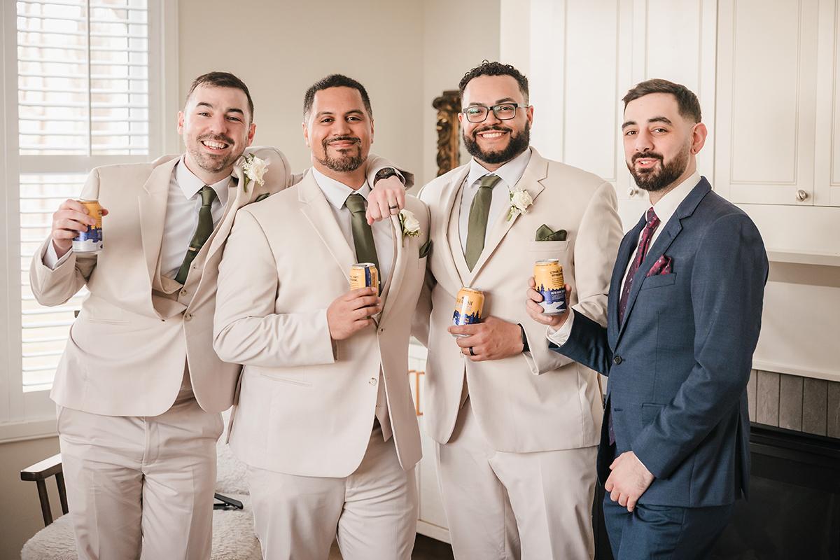 Groomsmen in suits holding canned drinks, smiling together indoors.