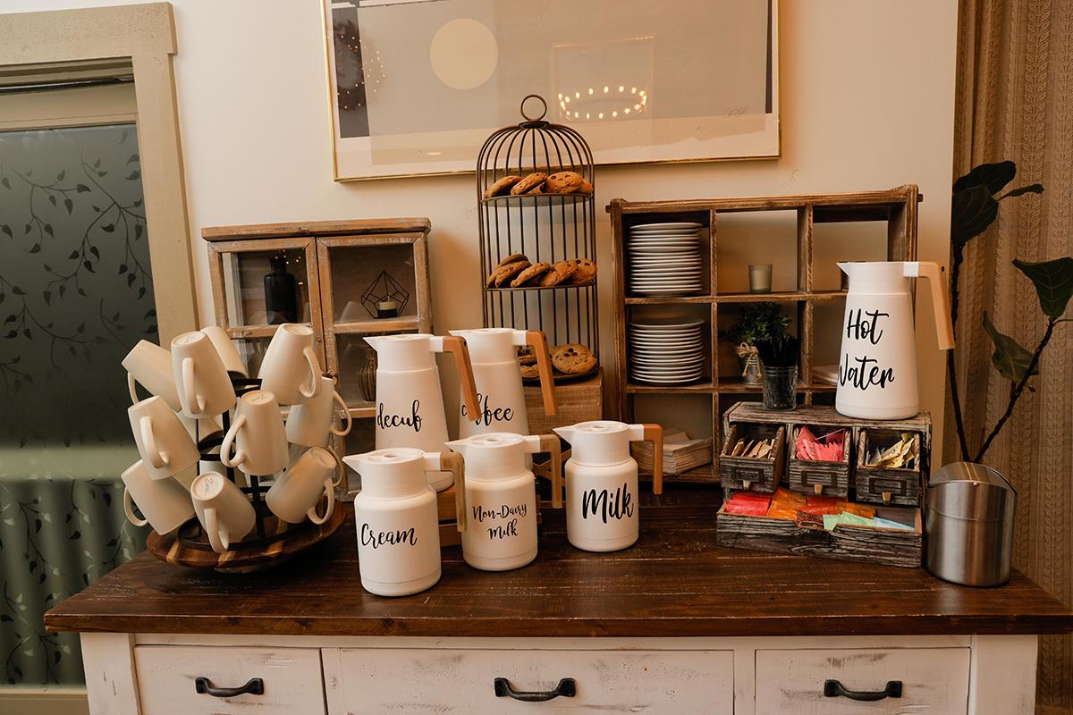 Rustic coffee bar with mugs, jars, pastries, and a “But first coffee” sign.