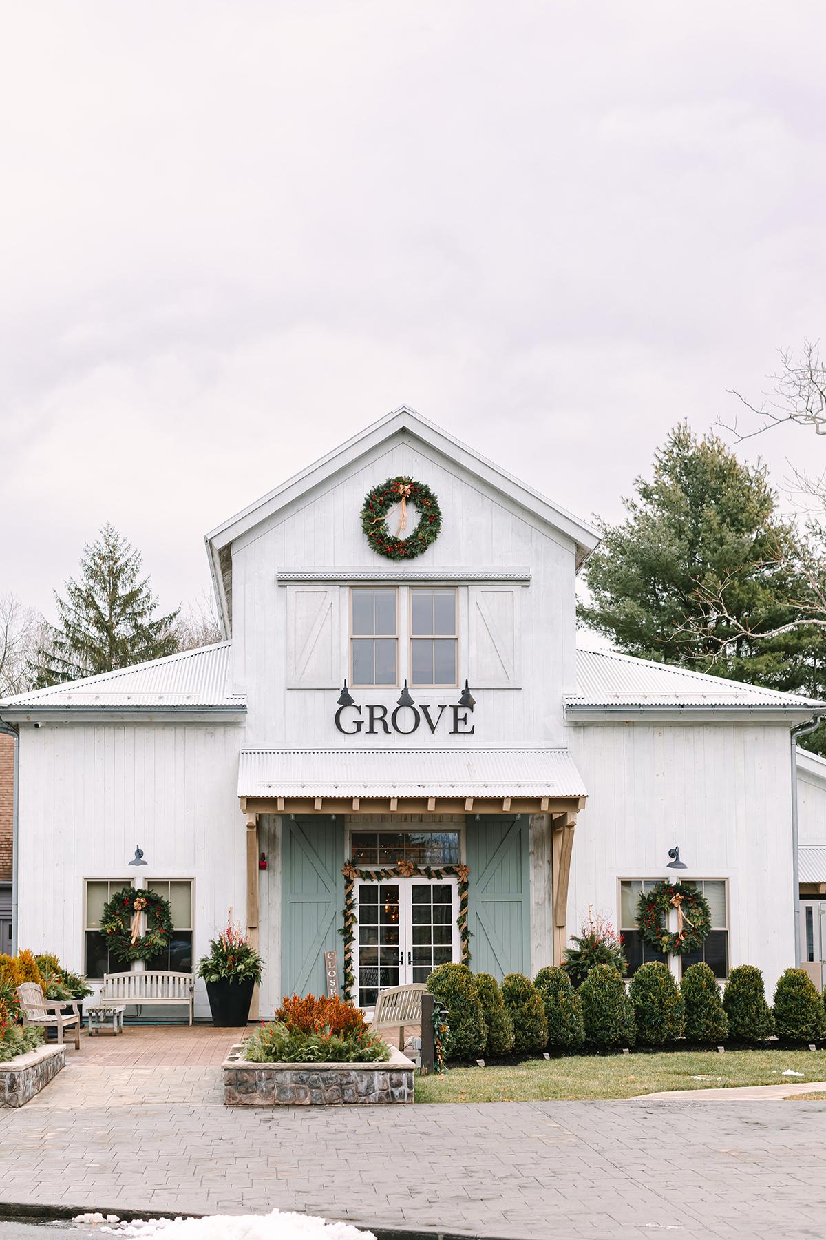 White farmhouse-style Grove storefront with wreath, shrubs, and autumn planters.
