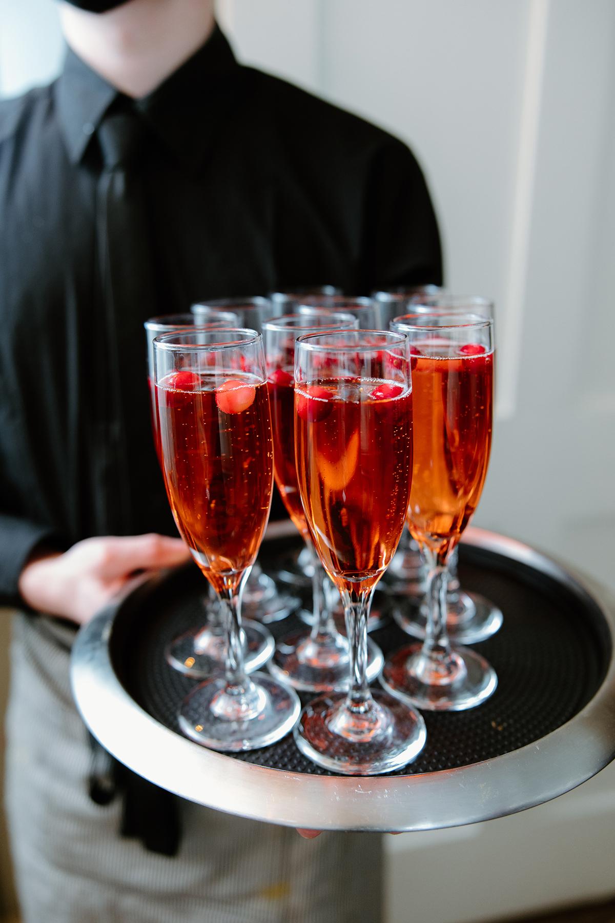 Server holding a tray of champagne flutes filled with red sparkling drinks.