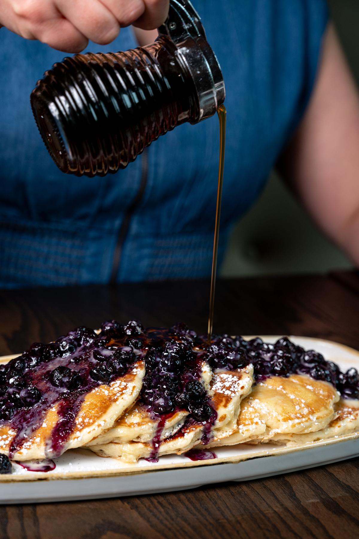 Person pouring dark syrup over pancakes topped with blueberries and cream
