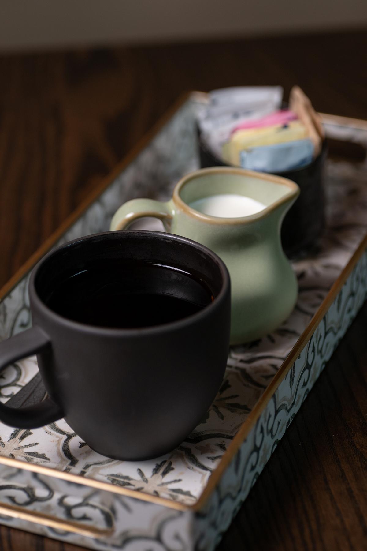 Black coffee mug and pale green creamer on a patterned tray.