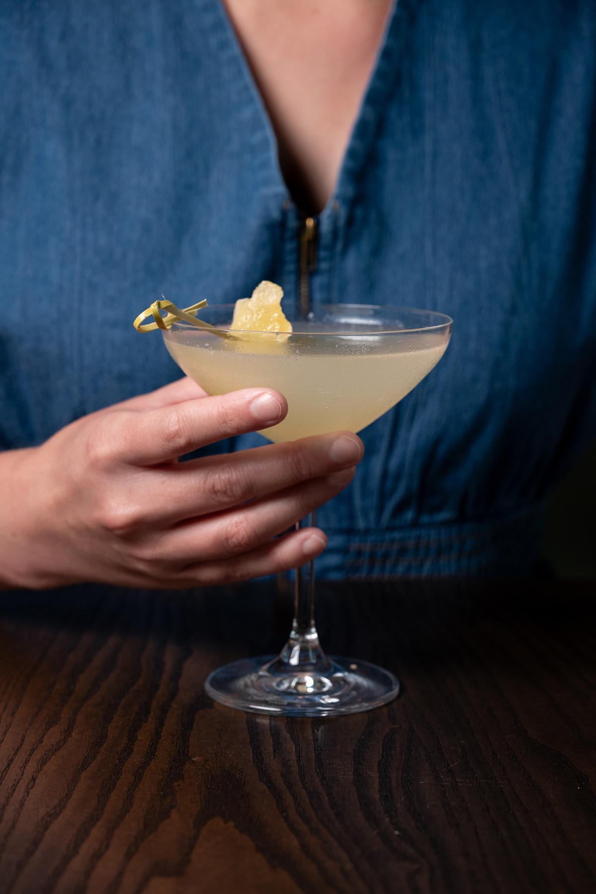 Person holding a pale cocktail in a coupe glass, garnished with lemon peel.