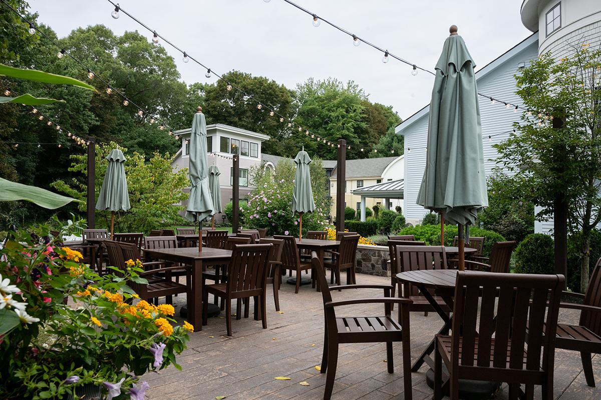 Outdoor restaurant patio with wooden tables, chairs, umbrellas, and flower beds.