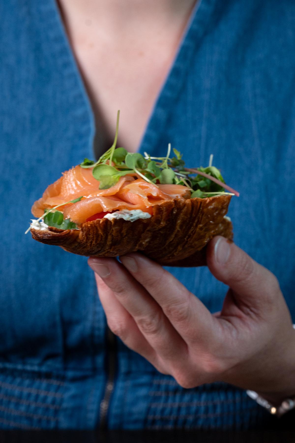Person in blue holding a rustic sandwich topped with greens and smoked salmon